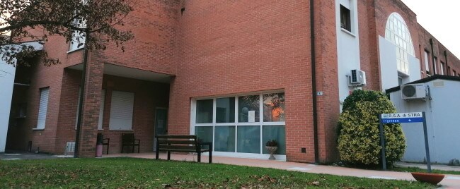 Ingresso edificio in mattoni con grandi finestre, panchine e piante, prato curato e insegna informativa